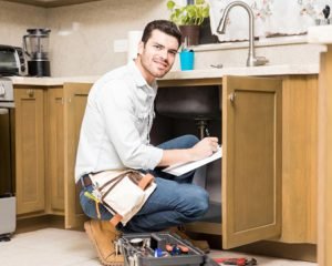 Plumber inspecting a kitchen sink and taking notes beside a toolbox